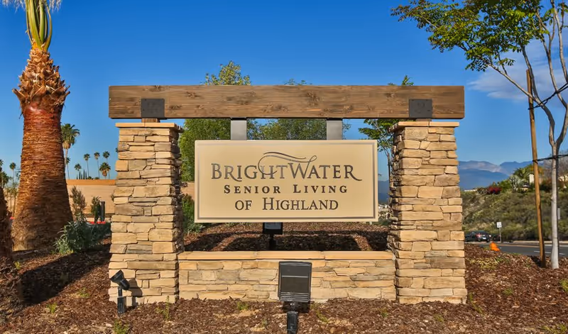 Stone and wood entrance sign for Brightwater Senior Living of Highland with landscaping and a clear blue sky in the background.
