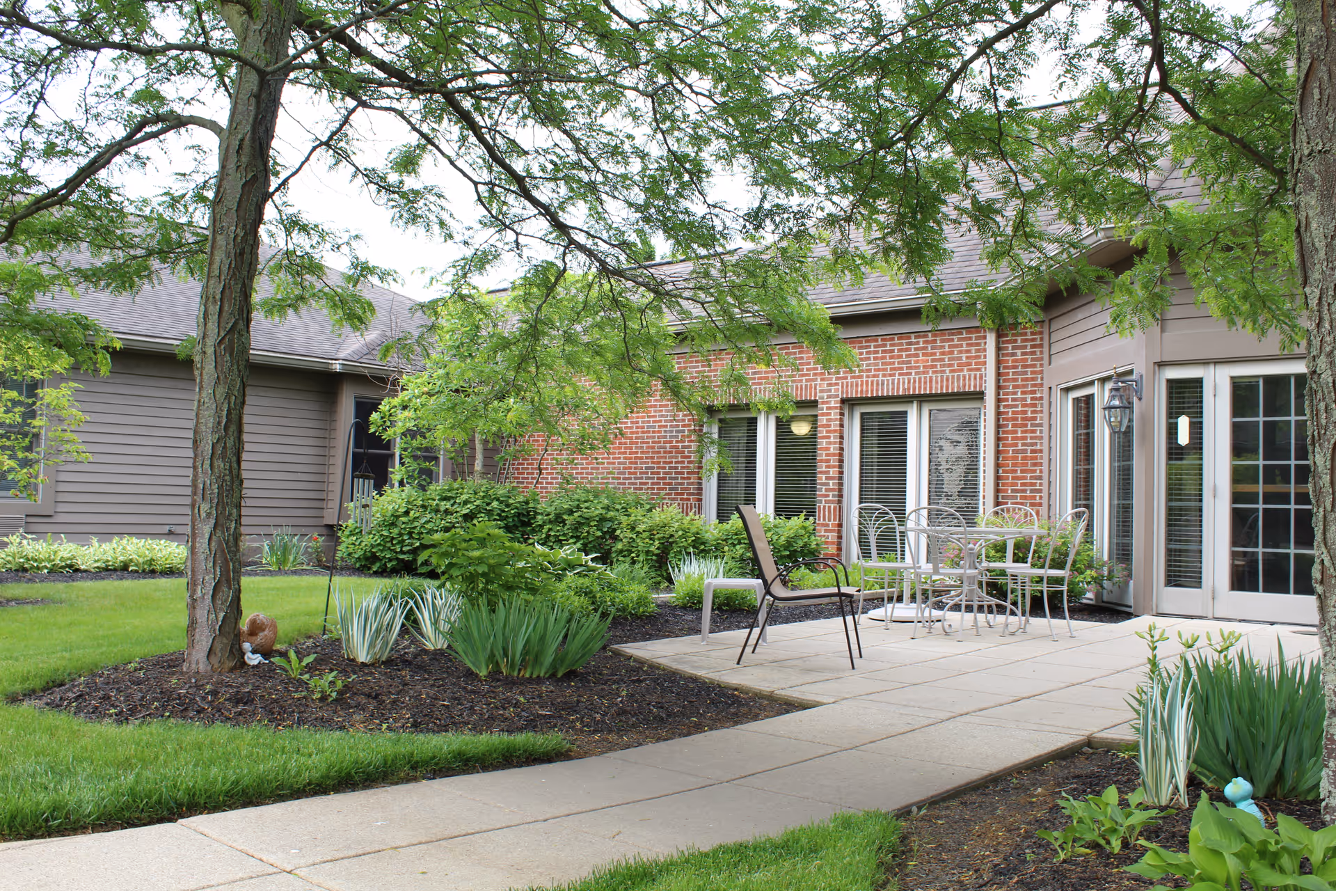 Outdoor patio area at Garbry Ridge featuring a paved walkway, metal table with chairs, a single lounge chair, surrounded by green plants, trees, and a brick and siding building in the background.