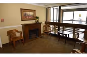 Small lobby seating area with wooden chairs, a bench, a framed painting above an electric fireplace, and a windowed entrance.