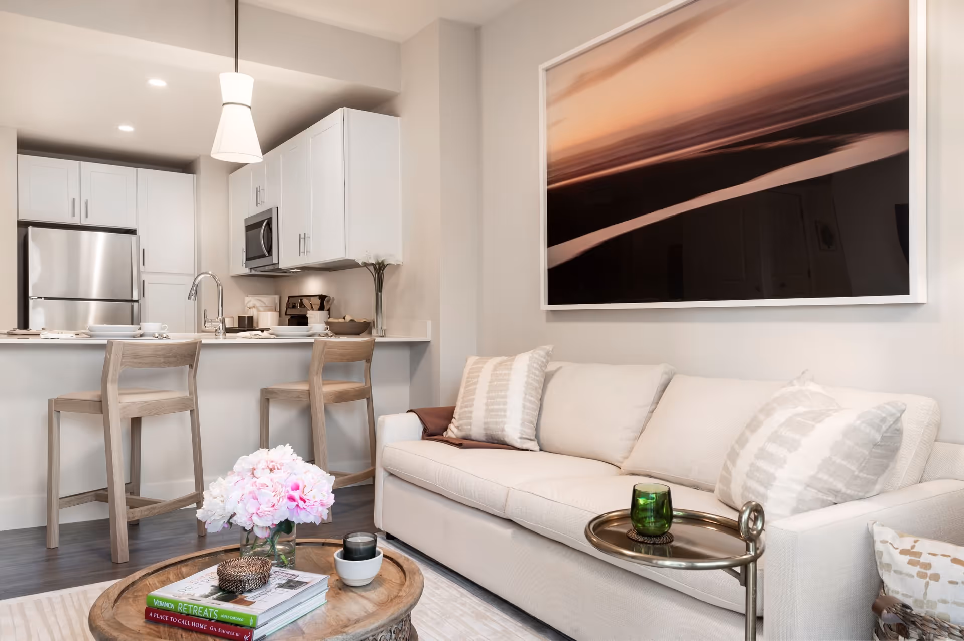 A modern living room and kitchen area featuring a beige sofa with patterned pillows, a round wooden coffee table with books and a flower vase, two wooden bar stools at a kitchen counter, white cabinetry, stainless steel refrigerator and microwave, and a large abstract wall art above the sofa.