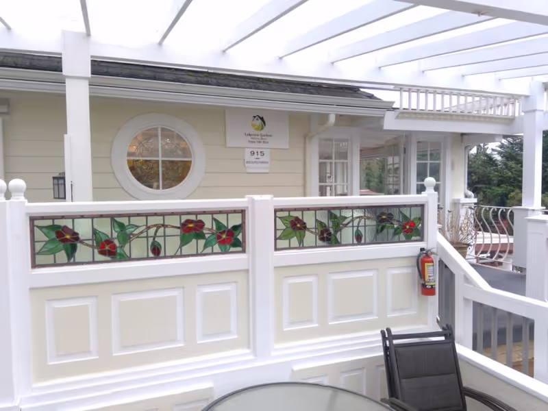 Outdoor patio area of Lakeview Gardens Adult Family Home featuring white wooden railings with decorative stained glass panels depicting red flowers and green leaves. There is a round glass table with a black chair in the foreground, a fire extinguisher mounted on the railing, and a beige building with a round window and a sign displaying the facility name and address in the background. The patio is covered with a white pergola.