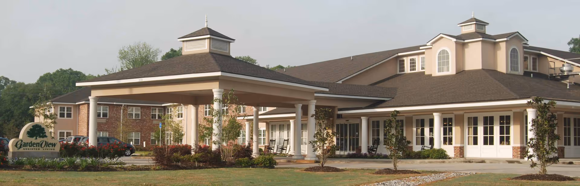 Exterior view of Garden View Assisted Living facility showing a large building with a covered entrance supported by white columns, surrounded by landscaped greenery and a parking area with cars.