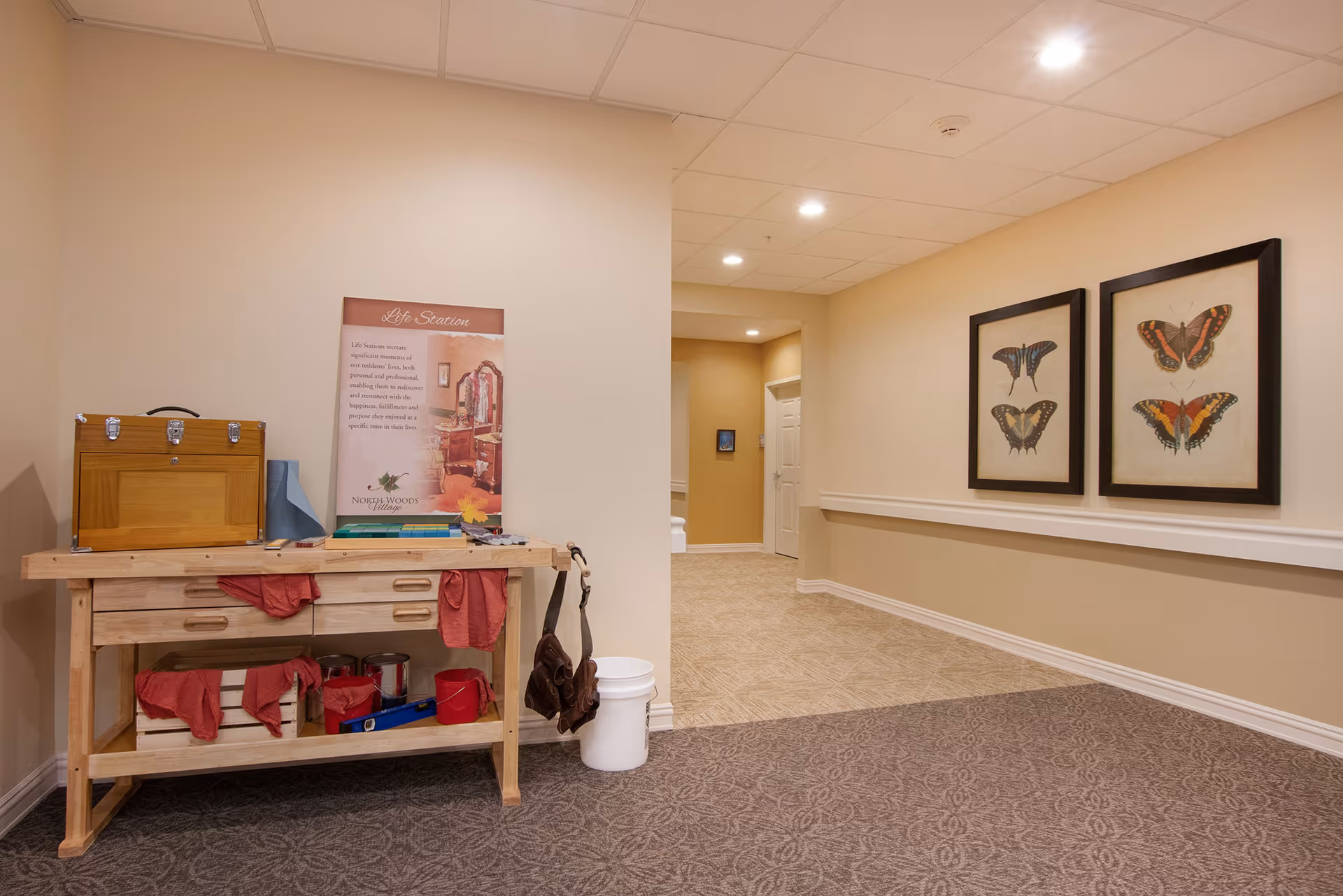 Interior hallway of North Woods Village of Kalamazoo with beige walls and ceiling lights. On the left side, there is a wooden workbench with drawers, paint cans, and red cloths. Above the workbench is a sign titled 'Life Station' with text and an image. On the right wall, there are two framed pictures of butterflies. The floor has two different carpet patterns, one in the foreground and one in the hallway.