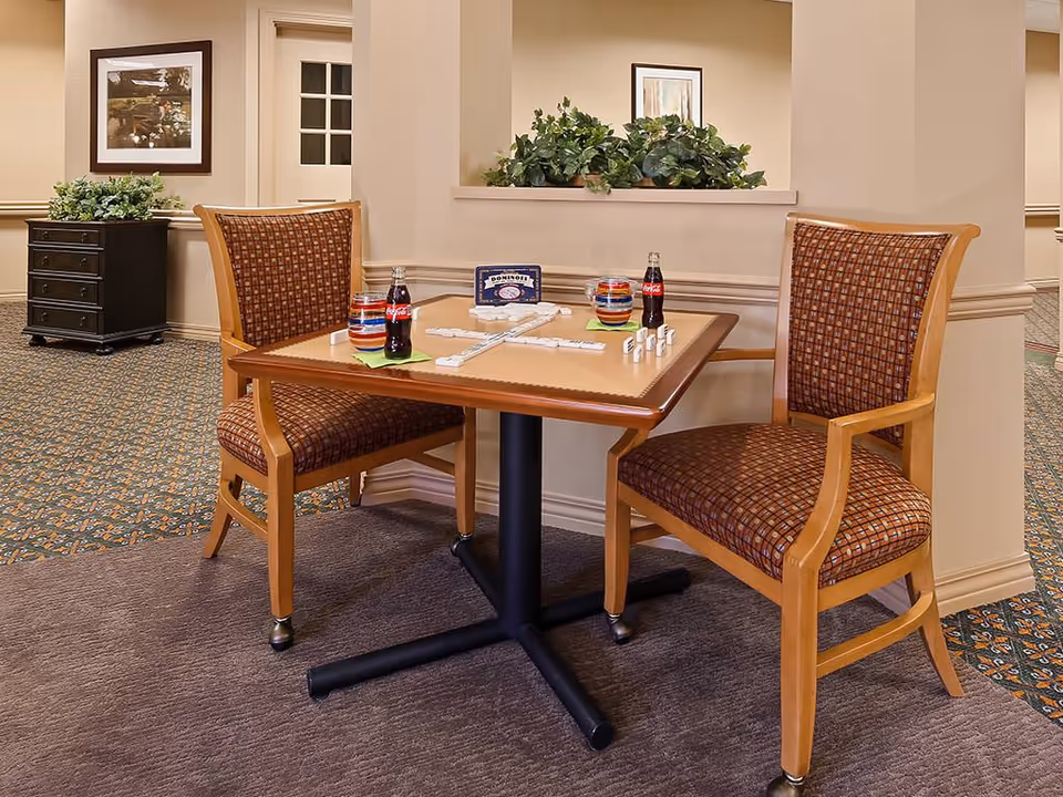 A small table with two cushioned wooden chairs in a carpeted room. On the table are two bottles of Coca-Cola, two colorful bowls, and a domino game set up with tiles. Behind the table is a half wall with green plants on top, and framed artwork is visible on the walls in the background.