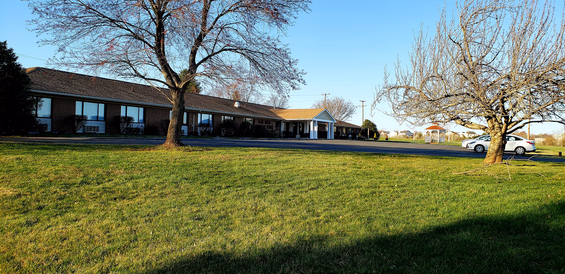 Exterior view of a single-story brick building with a covered entrance, surrounded by a grassy lawn and leafless trees under a clear blue sky. A white car is parked near the building, and a small gazebo is visible in the background.