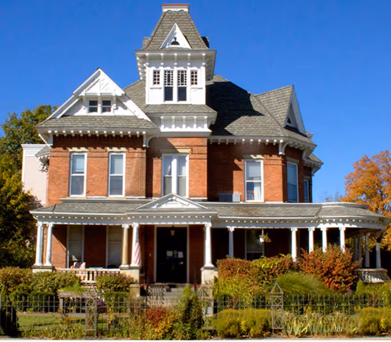 A large, historic brick building with white trim and a steep, multi-gabled roof under a clear blue sky. The building has a wrap-around porch with columns and is surrounded by green shrubs and a small garden with a metal fence in front.