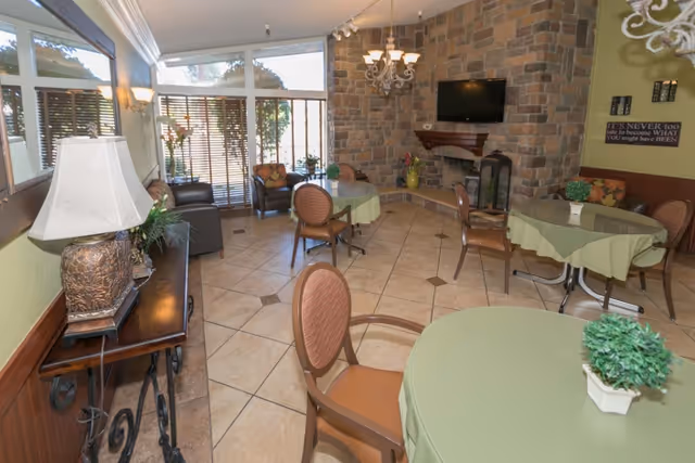 Bright communal dining/lounge area with round tables draped in green tablecloths, chairs, a stone fireplace with a mounted TV, and large windows.