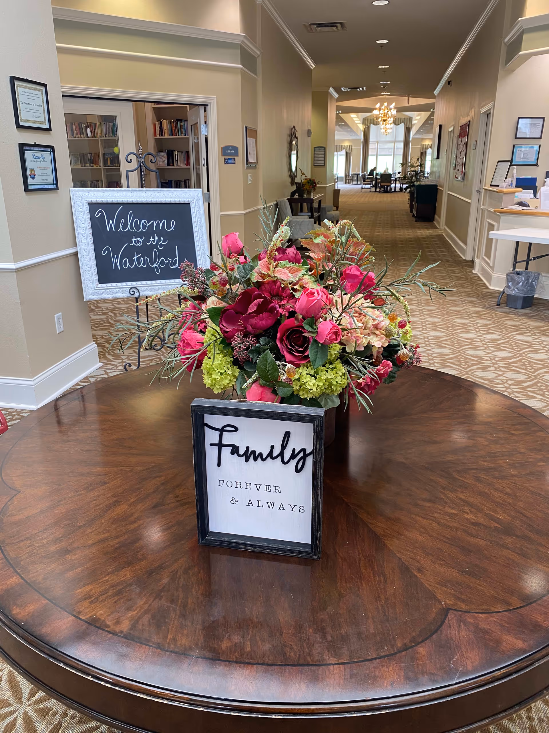 A round wooden table with a floral arrangement of pink and green flowers and a framed sign that reads 'Family FOREVER & ALWAYS' in the foreground. In the background, a hallway with carpeted floor and beige walls leads to a well-lit common area with chairs and tables. A chalkboard sign on an easel reads 'Welcome to the Waterford'.
