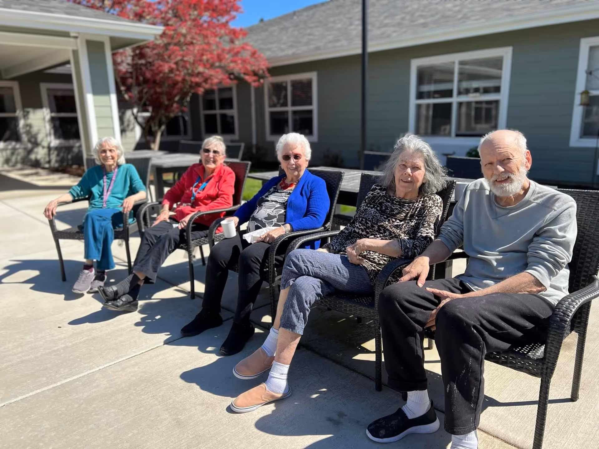 Five elderly individuals sitting outside on black wicker chairs in a sunny courtyard area with a building and a tree with red leaves in the background. They are smiling and appear to be enjoying the outdoor setting.