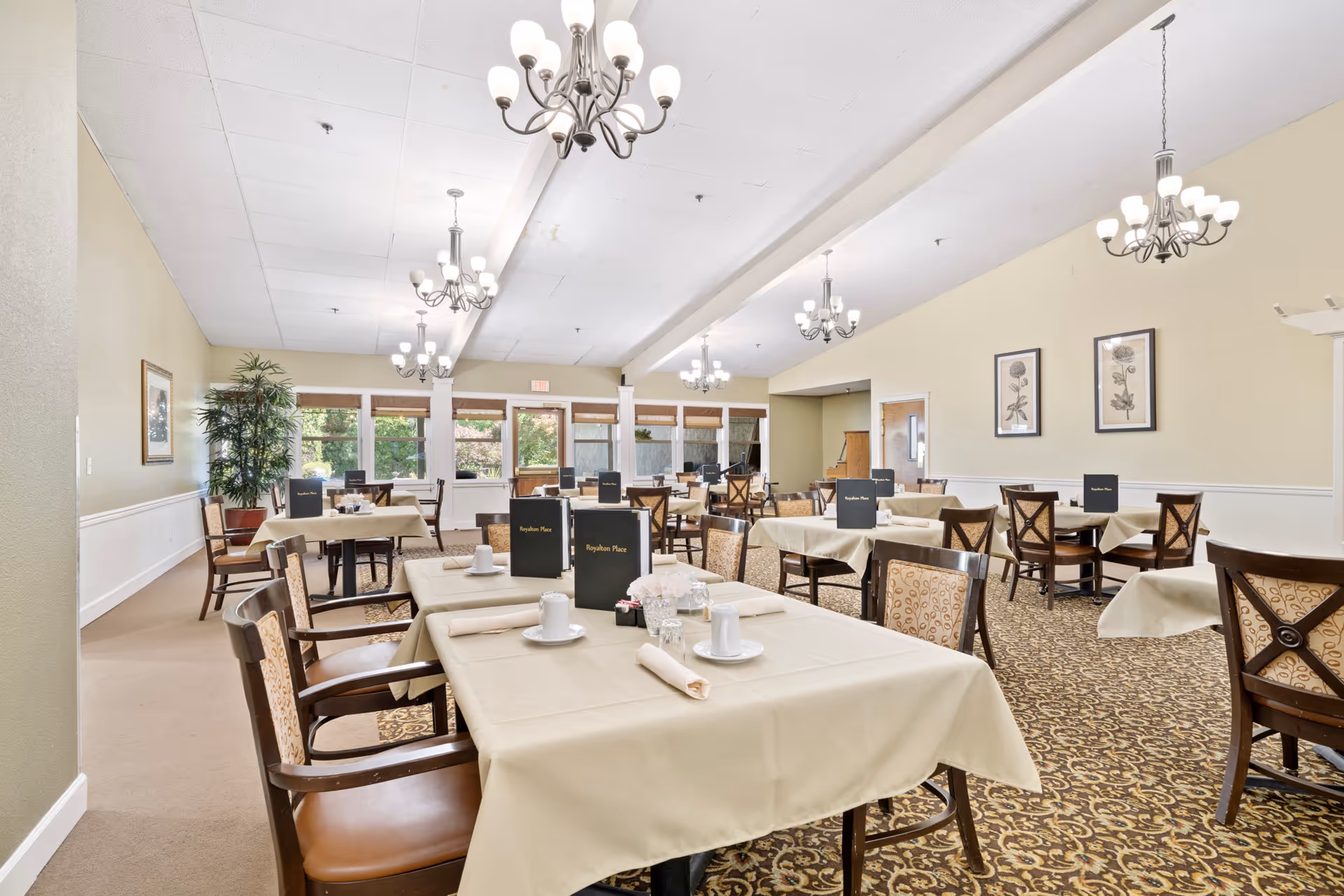 A spacious dining room with multiple tables covered in beige tablecloths, each set with white cups, plates, and napkins. Dark wooden chairs with patterned upholstery surround the tables. Several black menus labeled 'Royalton Place' are placed on the tables. The room features large windows letting in natural light, beige walls with framed botanical prints, and multiple chandeliers hanging from the ceiling. A large potted plant is visible in the corner.