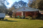 Exterior view of a single-story brick building with a triangular roof and a sign on the lawn in front. The building is surrounded by grass, small bushes, and trees, with a clear sky above.