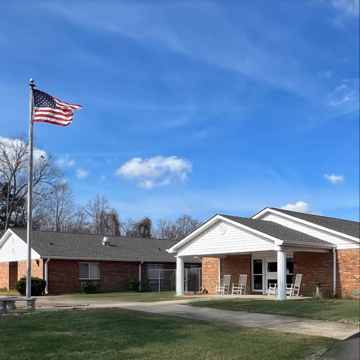 American flag flies beside a brick senior care building with a covered entrance and rocking chairs.