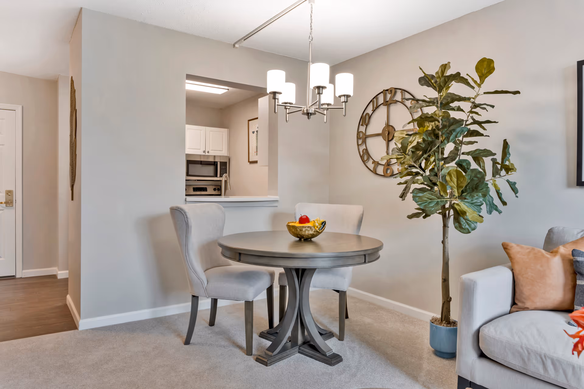 Cozy apartment dining area with a round table, two upholstered chairs, a potted plant, wall clock, and views into the kitchen and living room.