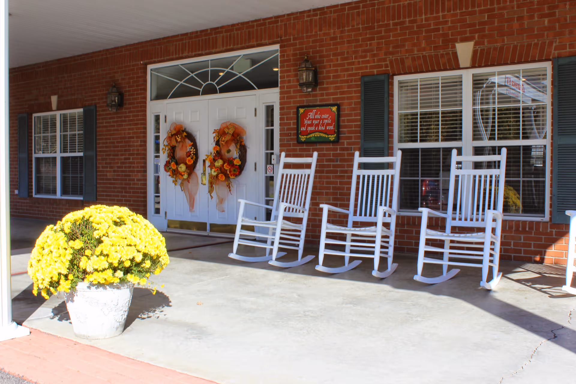 Covered brick entrance of a senior living community with white double doors decorated with wreaths, a row of white rocking chairs on the porch and a large pot of yellow flowers.