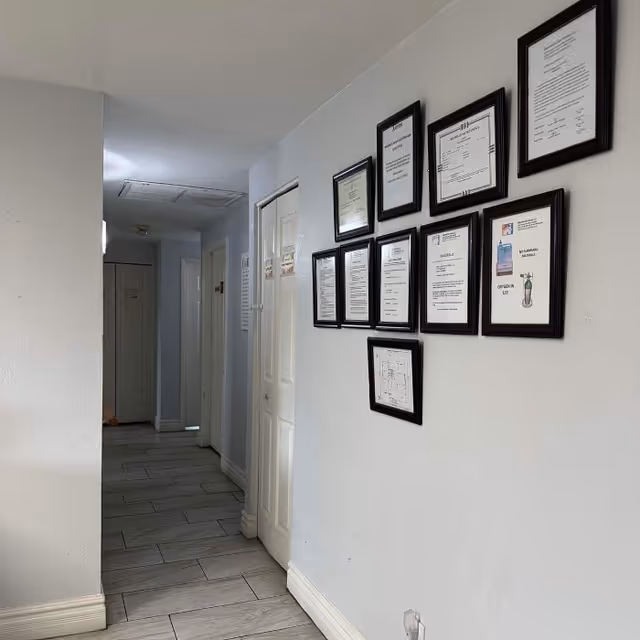 A hallway with light gray walls and tiled floor. On the right wall, there are multiple framed certificates and documents arranged in a grid pattern. Several doors are visible along the hallway.