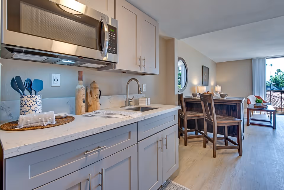 Modern kitchen area with gray cabinets, a white marble countertop, a stainless steel microwave, and a sink. On the counter are kitchen utensils in a decorative holder, two wooden bottles, and a folded towel. In the background, there is a dining area with a wooden table and chairs, a round window, and a living space with a small table and a large window with vertical blinds letting in natural light.