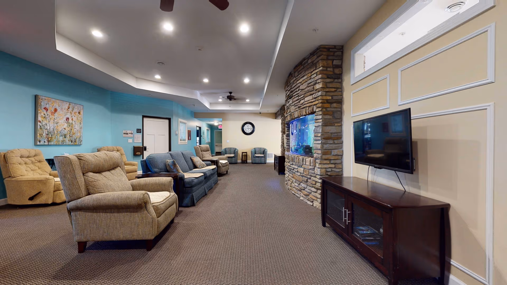 A spacious and well-lit living room area in a senior living facility featuring multiple comfortable armchairs and sofas arranged along the walls. The room has a stone accent wall with a built-in aquarium and a flat-screen TV mounted on the opposite wall above a wooden cabinet. The walls are painted in soft blue and beige tones, with framed artwork and a clock visible in the background.