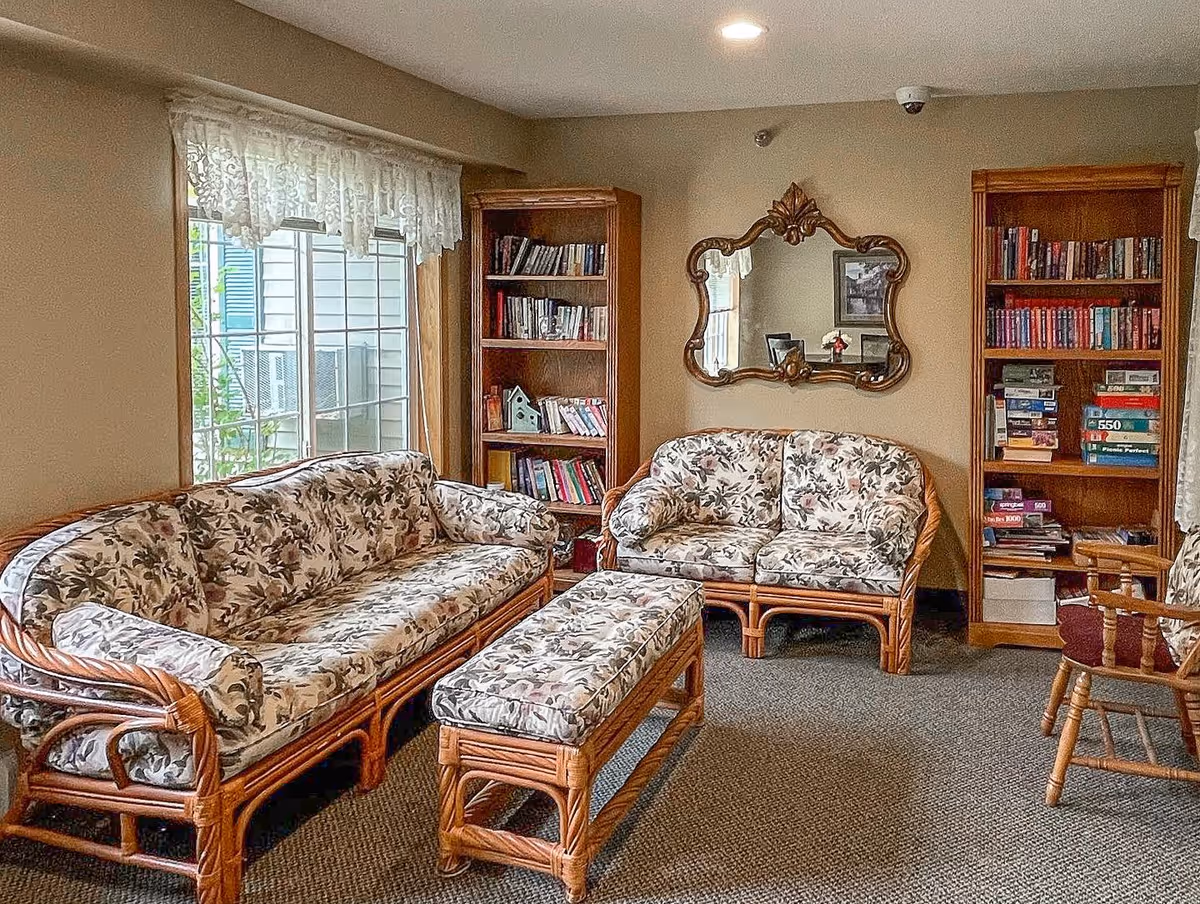 A cozy living room with floral-patterned cushioned wicker furniture including a sofa, loveseat, and an ottoman. Two wooden bookshelves filled with books and games flank a decorative wall mirror. A window with lace curtains lets in natural light, and a wooden chair is partially visible on the right.
