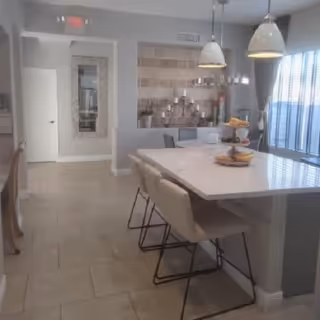 Interior view of a dining area with a large white marble table surrounded by beige cushioned chairs with black metal legs. Two pendant lights hang above the table. In the background, there is a wall with decorative tiles and a candelabra with multiple candles. A window with vertical blinds allows natural light into the room.