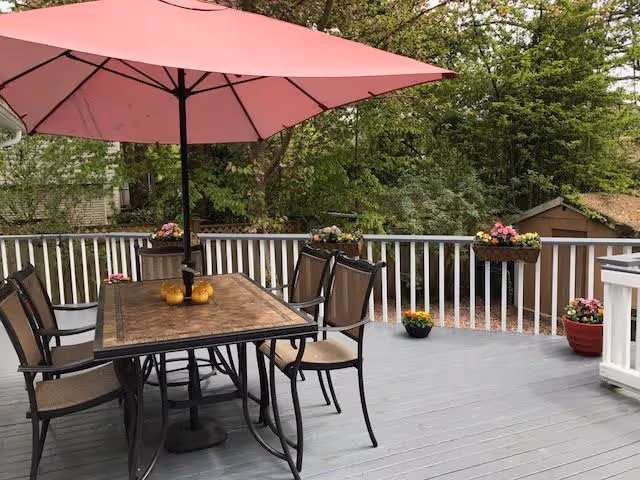 A wooden deck patio with a dining table, chairs and a pink umbrella surrounded by potted flowers and trees.