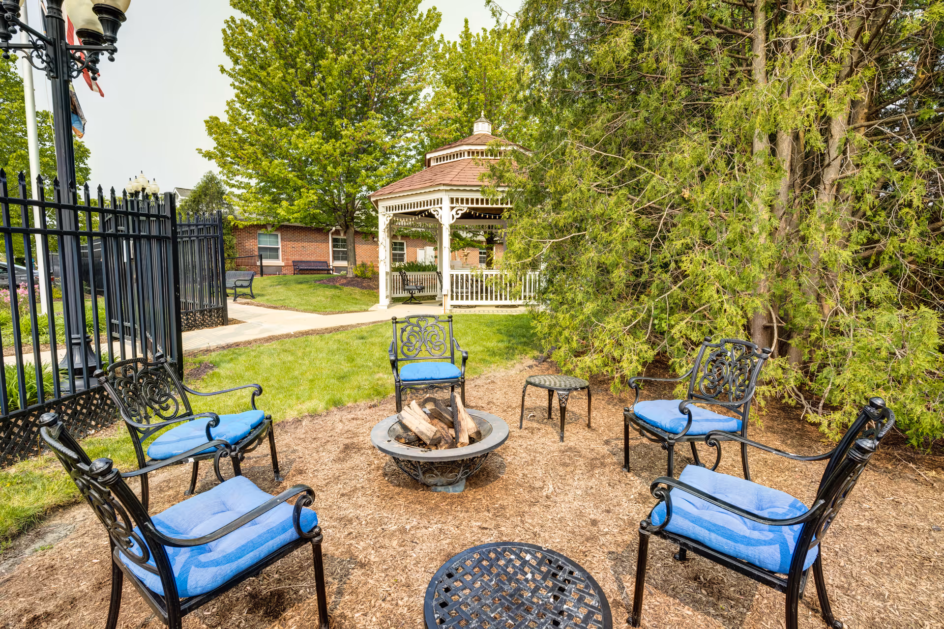 Outdoor seating area with five black metal chairs with blue cushions arranged around a fire pit. There is a small black metal table nearby. In the background, there is a white gazebo surrounded by green trees and grass, with a brick building partially visible behind the gazebo.