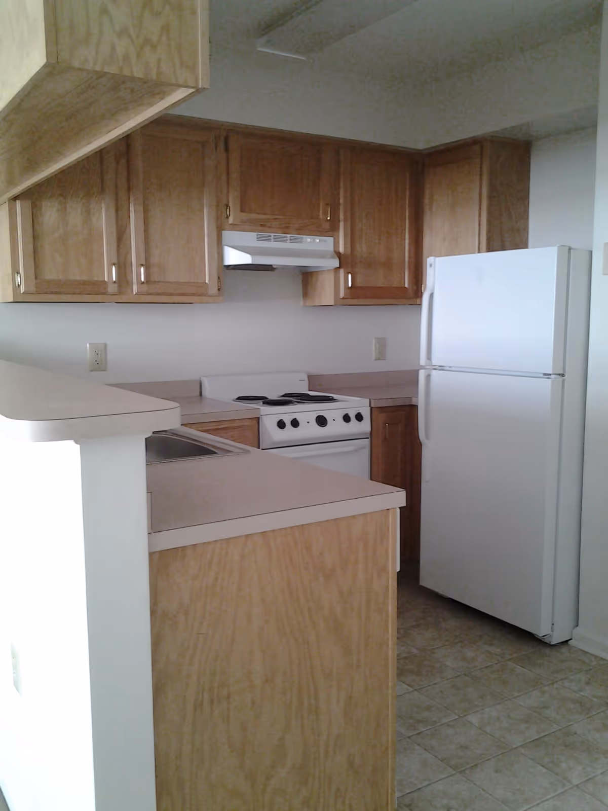 A small kitchen with light wood cabinets, a white refrigerator, a white stove with an exhaust hood, and a countertop with a sink. The floor is tiled with light-colored tiles.