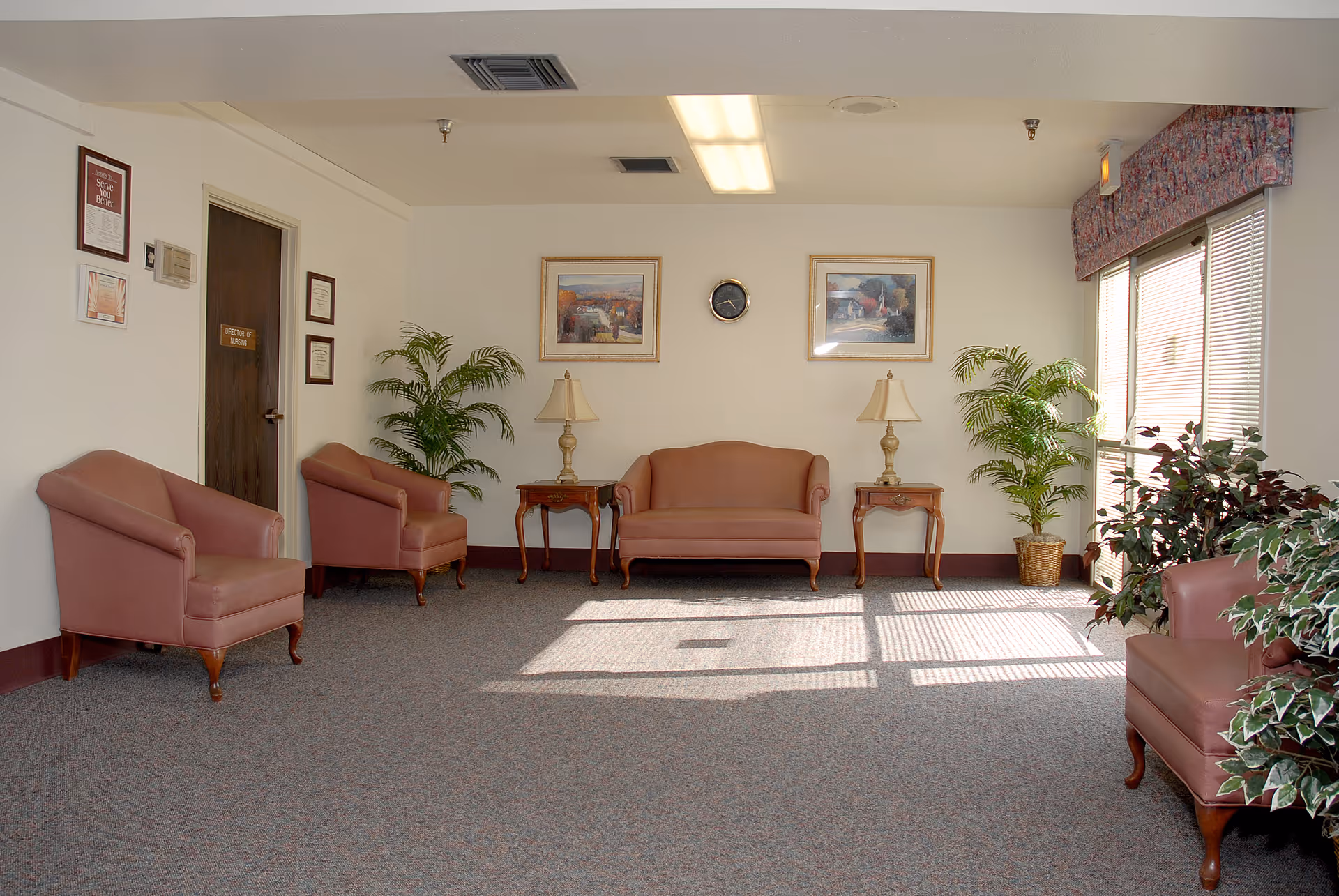 A well-lit waiting area with several pink upholstered chairs and a matching loveseat arranged around two wooden side tables with lamps. The room has carpeted flooring, two framed paintings on the wall, a clock, and several potted plants near a large window with blinds and a floral valance.