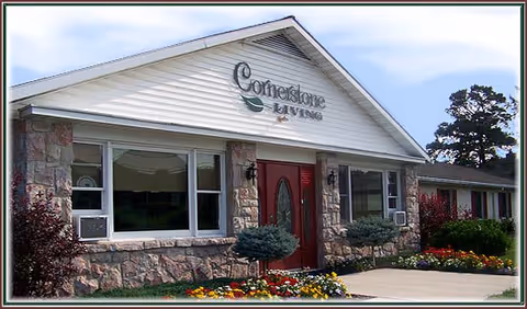 Front exterior view of Cornerstone Living facility showing a single-story building with stone and white siding, a red door, large windows, and landscaped flower beds in front.