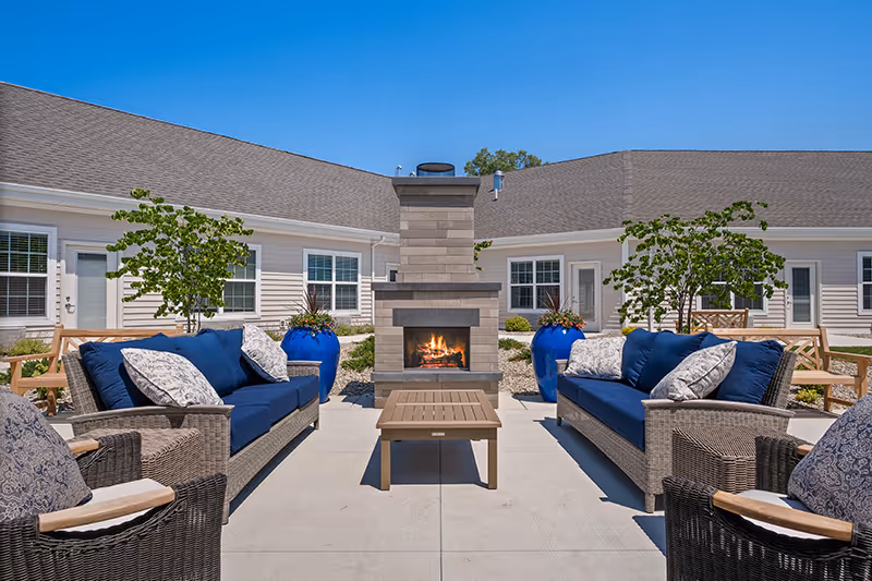 Outdoor patio area at Quincy Place Senior Living featuring two blue cushioned sofas, two wicker armchairs with patterned cushions, a central wooden coffee table, and a stone fireplace with a fire burning. The patio is surrounded by beige buildings with white trim, windows, doors, and some greenery including small trees and large blue planters.