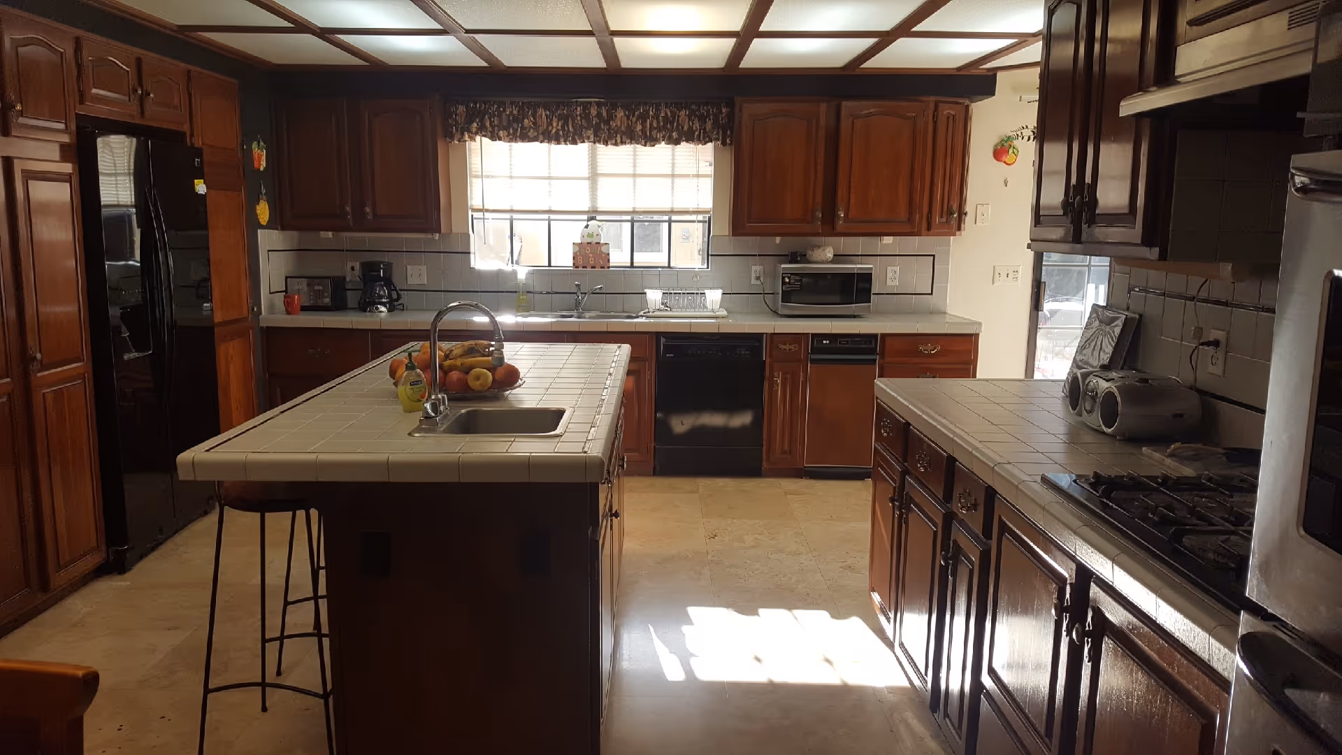 Large kitchen with a central tiled island and sink, wooden cabinets, and appliances under a window.