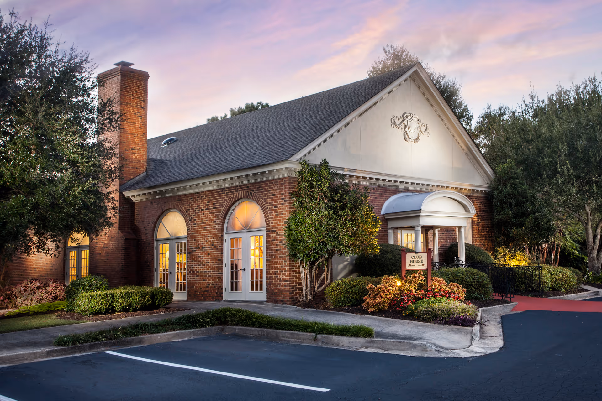 Exterior view of a brick building with large arched windows and a chimney, surrounded by landscaped bushes and trees, with a sign reading 'Club House' near the entrance, under a colorful evening sky.