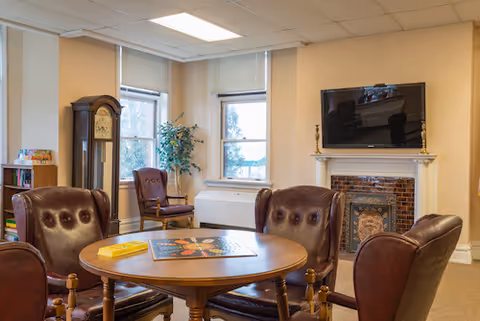 A cozy common area with four brown leather chairs arranged around a round wooden table with a board game on it. The room has two large windows with blinds, a tall grandfather clock, a potted plant, a fireplace with a decorative tile surround, and a flat-screen TV mounted above the fireplace.