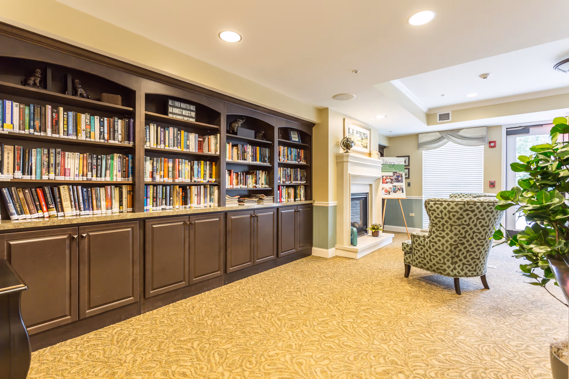 A cozy library and sitting area in an assisted living facility featuring a long dark wood bookshelf filled with books, a fireplace with decorative items on the mantel, a patterned armchair, a large window with blinds, and a potted plant near the entrance door.