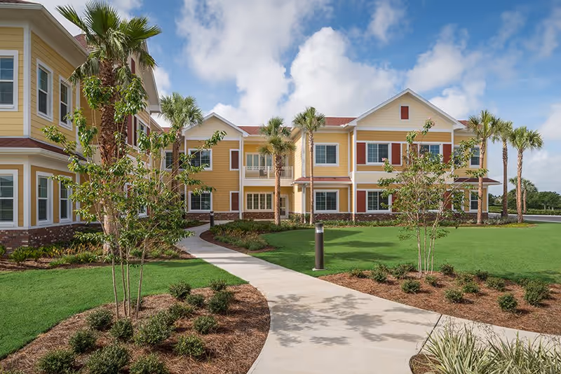 Exterior view of a two-story assisted living facility with yellow siding, red shutters, and a red roof. The building is surrounded by a well-maintained lawn, palm trees, and landscaped bushes. A curved concrete walkway leads to the entrance under a partly cloudy sky.