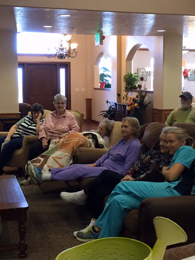 A group of senior residents seated on sofas in a communal living room area with a coffee table and decorative plants.