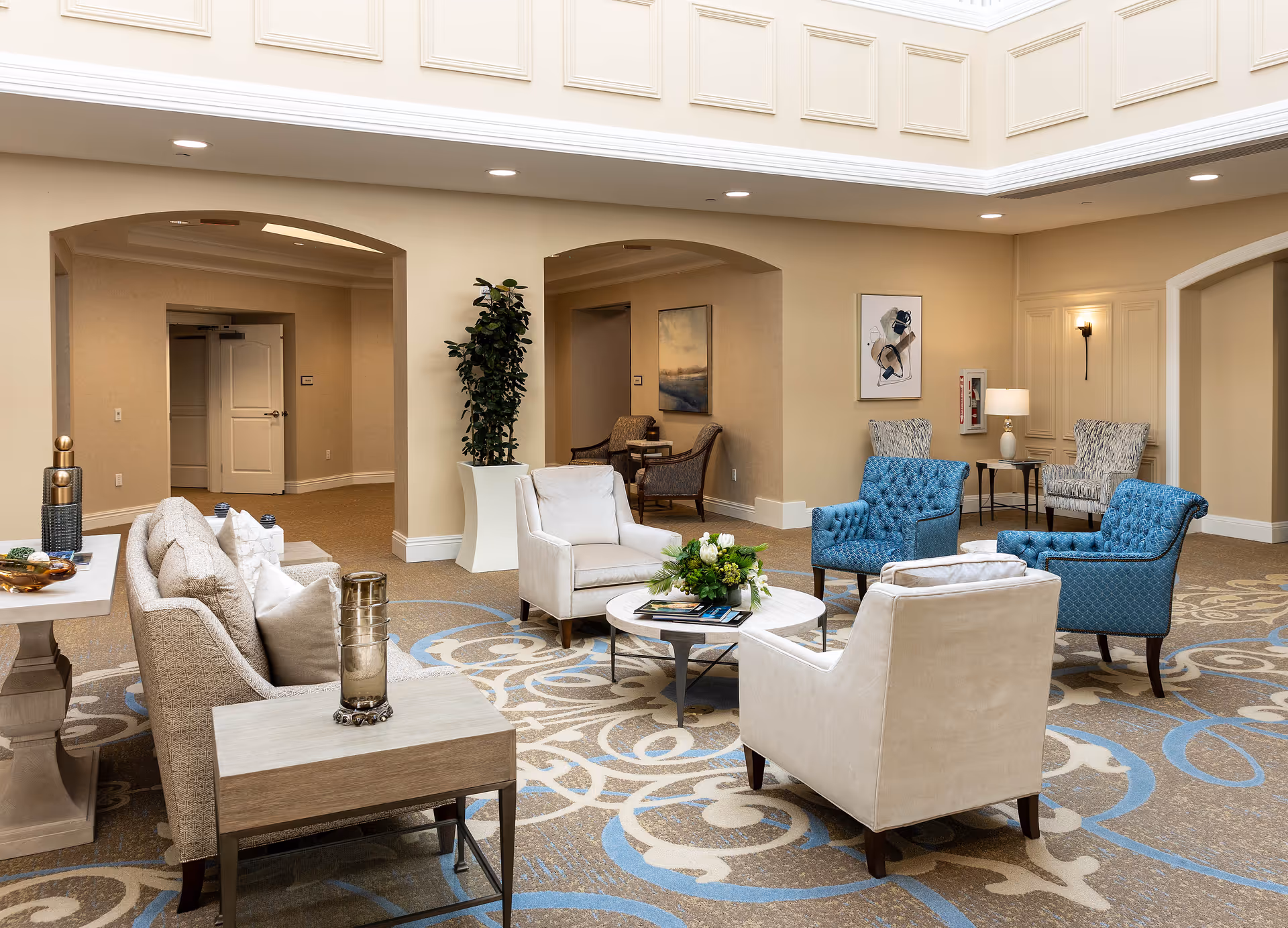 A spacious and well-lit common area in a retirement community featuring a seating arrangement with beige and blue upholstered chairs around a round coffee table with a floral centerpiece. The room has beige walls, decorative molding near the ceiling, and a patterned carpet with blue and beige swirls. There are two archways leading to other rooms, a tall potted plant, framed artwork on the walls, and a side table with decorative items.