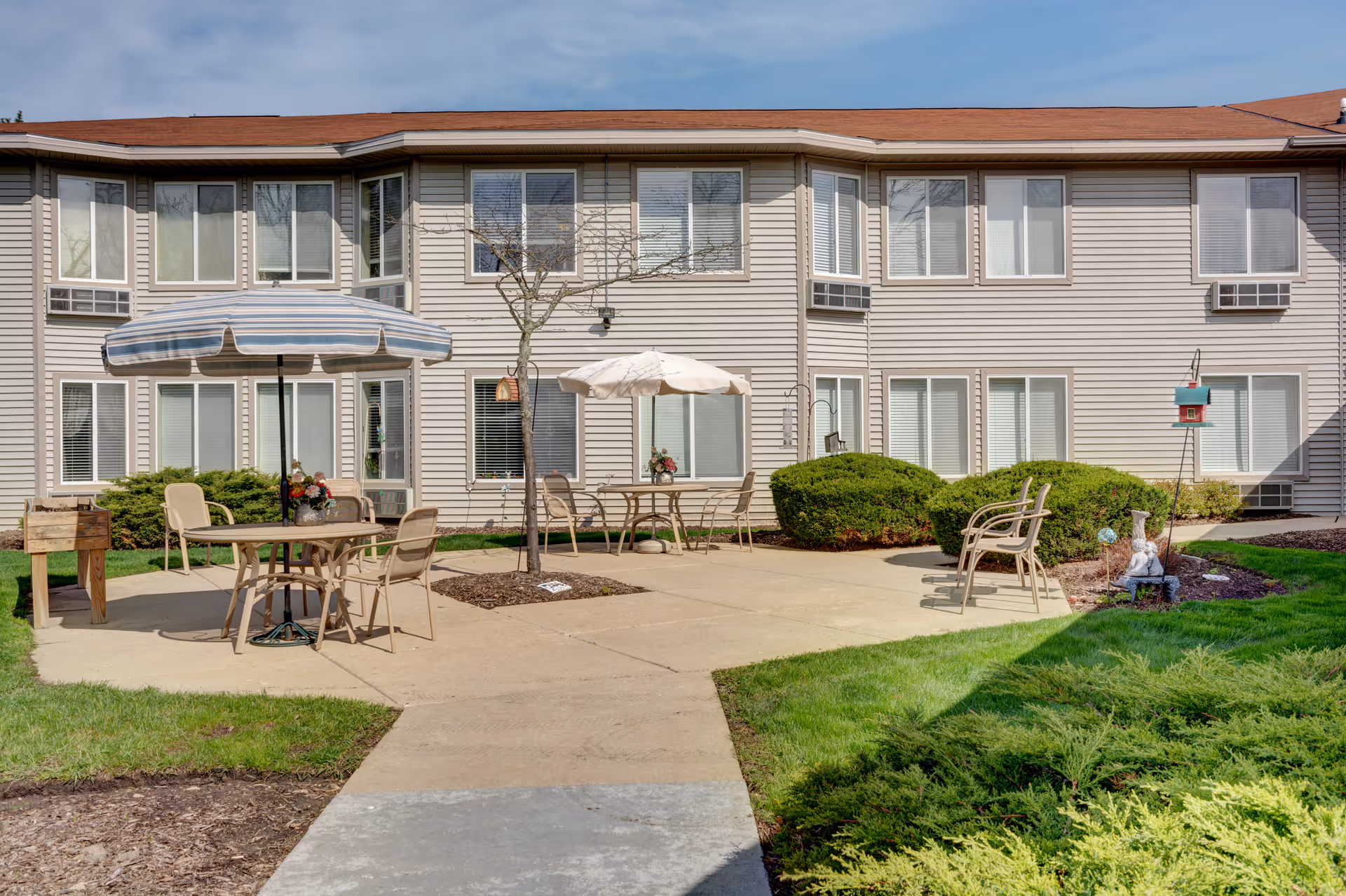 Sunlit courtyard with patio tables, umbrellas, chairs, and landscaping in front of a two-story residential building.