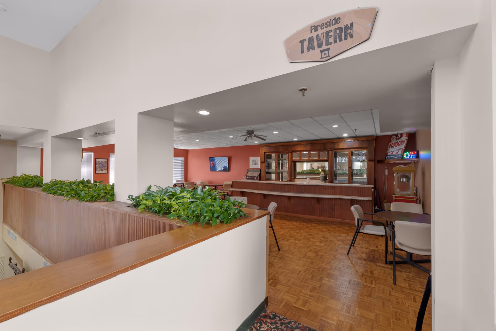 Interior view of a retirement community lounge area named Fireside Tavern, featuring a wooden bar counter, tables with chairs, a popcorn machine, and a television mounted on a red wall.
