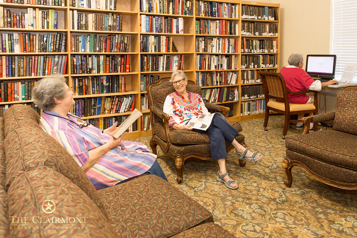 A cozy library room in a retirement community with bookshelves filled with books along the wall. Two elderly women are seated on upholstered chairs and a sofa, reading and chatting. Another elderly woman is seated at a desk using a computer near a window with blinds.