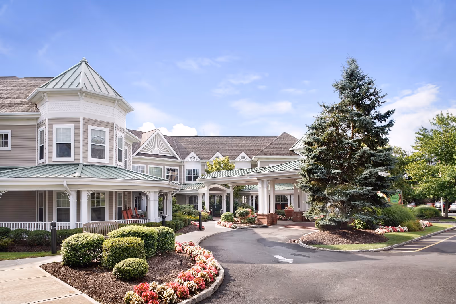 Exterior view of Sunrise of Woodcliff Lake senior living facility showing a large building with a covered entrance, landscaped bushes, flowers, and a tall evergreen tree under a blue sky with some clouds.