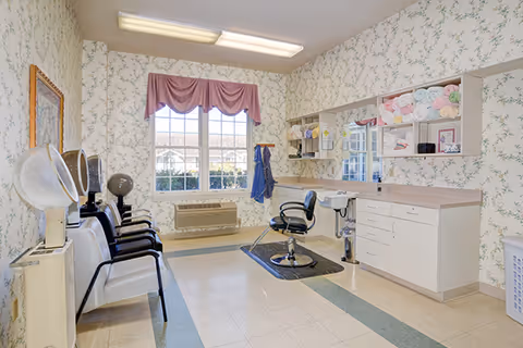 A bright hair salon room with floral wallpaper, a large window with a pink valance, three white chairs with black armrests lined up next to hair drying machines, a black salon chair in front of a counter with a large mirror, and shelves holding rolled towels and hair care products.