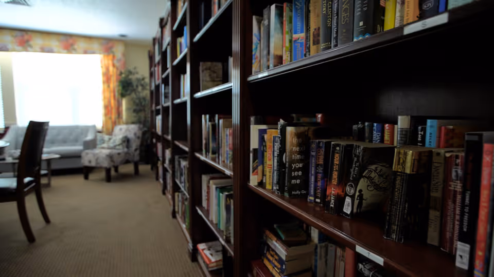 A wooden bookshelf filled with books in a cozy lounge with chairs and a sofa by a bright window.