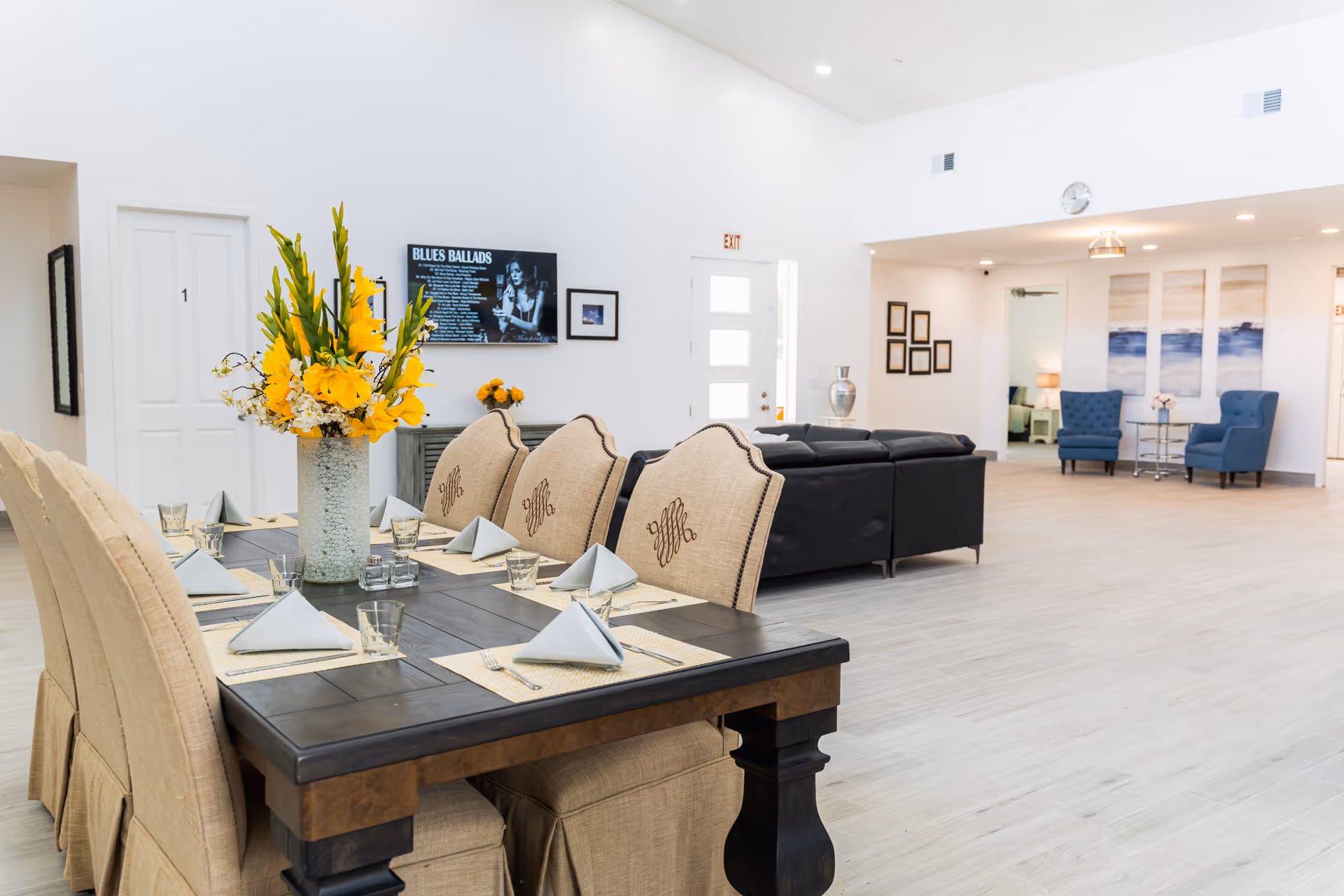 A bright and spacious interior room featuring a dining table set with beige upholstered chairs, napkins, and glasses. A vase with yellow flowers is placed in the center of the table. In the background, there is a black leather sofa, two blue armchairs, framed pictures on the walls, and a TV screen displaying 'BLUES BALLADS'. The room has light-colored wooden flooring and white walls with recessed lighting.