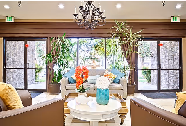 A bright and welcoming living room area with large glass doors and windows letting in natural light. The room features a beige sofa with colorful cushions, two brown armchairs with yellow pillows, a round white coffee table with decorative flowers, and several tall green plants. A chandelier hangs from the ceiling, and exit signs are visible above the doors.
