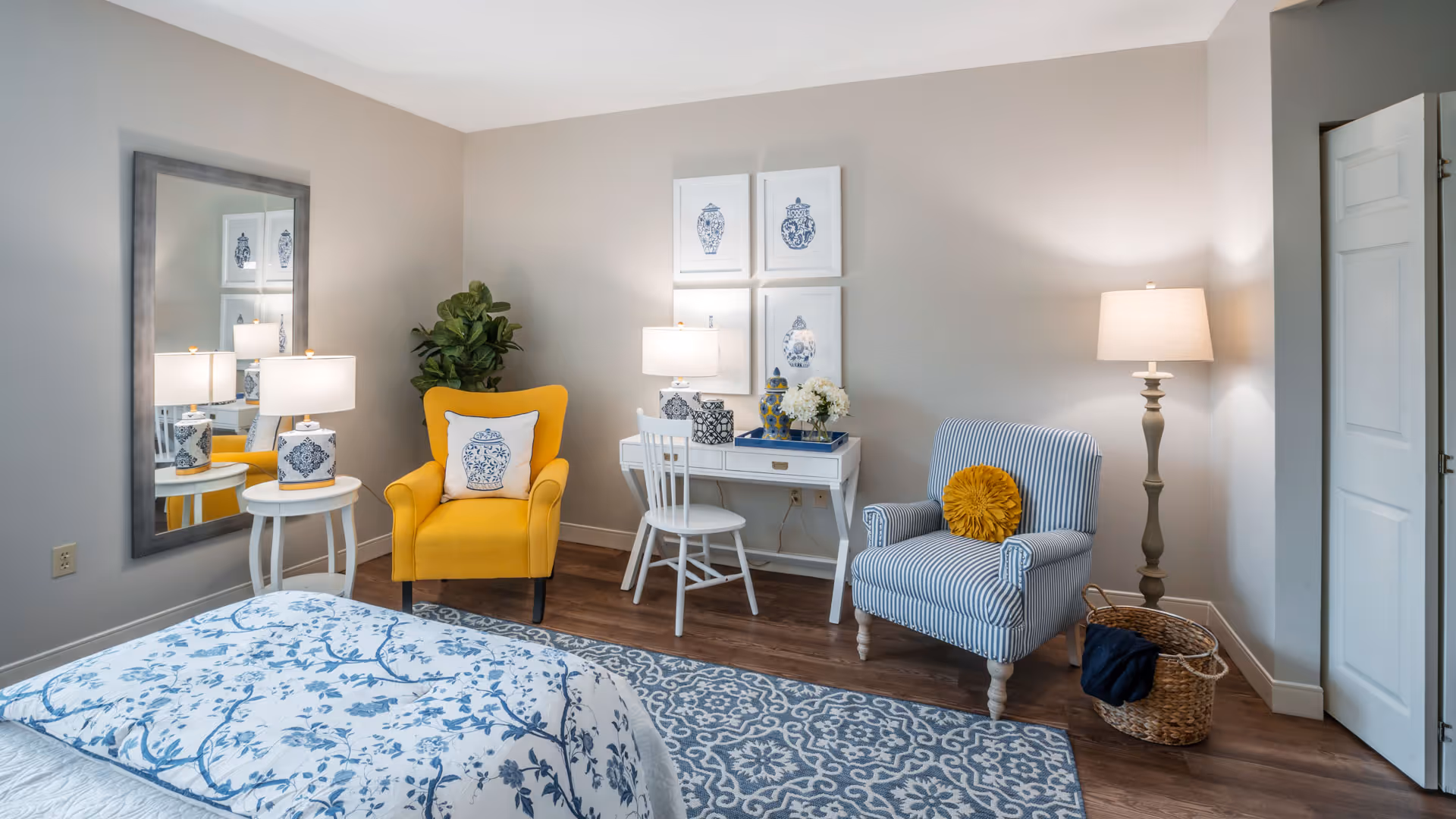 A cozy bedroom featuring a bed with blue and white floral bedding, a yellow armchair with a decorative pillow, a white side table with a lamp, a large wall mirror, a small white desk with a chair and decorative items, a blue and white striped armchair with a yellow round pillow, a floor lamp, a woven basket, and framed blue and white artwork on the wall.