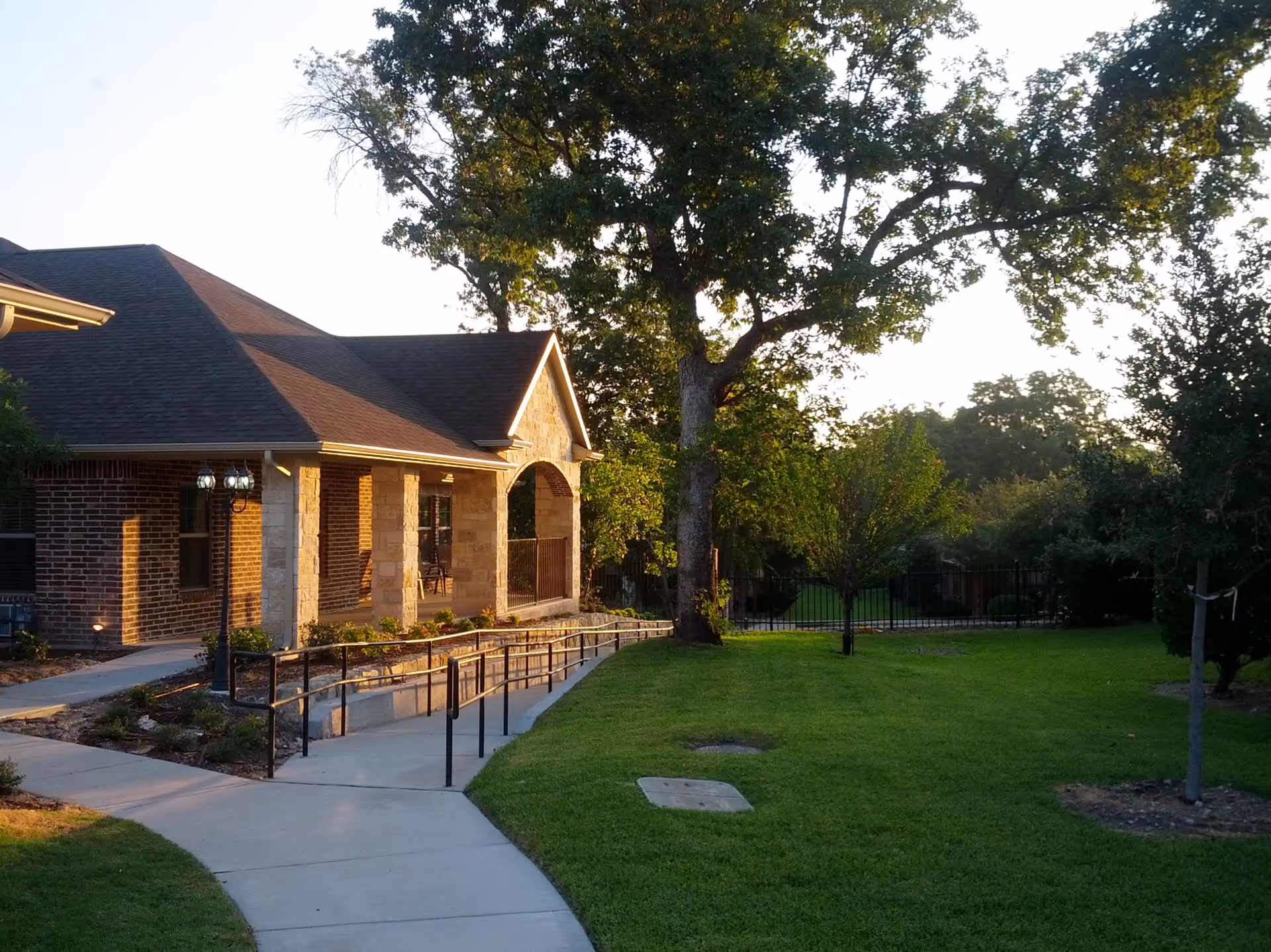 Single-story brick assisted living building with a covered porch, wheelchair ramp, and a grassy yard with large trees at sunset.