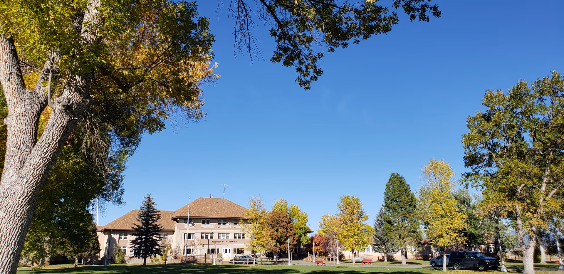 Exterior view of the Masonic Home of Montana building surrounded by green grass, trees with autumn foliage, and a clear blue sky.