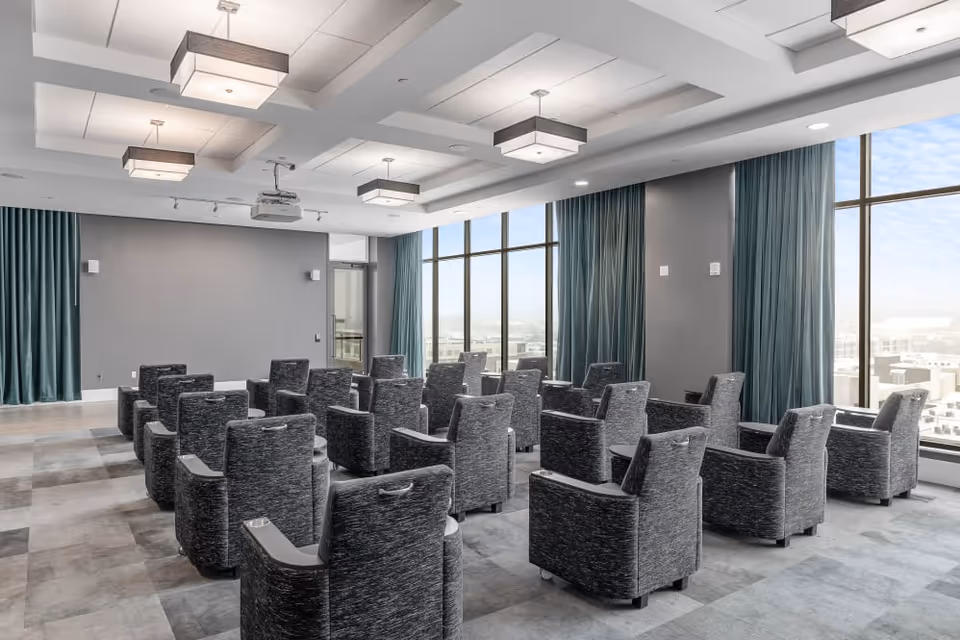 A modern meeting or presentation room with multiple rows of gray upholstered chairs facing a blank gray wall with a projector mounted on the ceiling. Large windows with teal curtains allow natural light to fill the space.