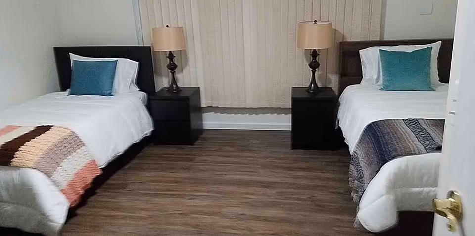A bedroom with two single beds placed parallel to each other, each with white bedding, a decorative pillow, and a knitted throw blanket. Between the beds are two black nightstands, each with a lamp. The room has wooden flooring and a window with vertical blinds in the background.
