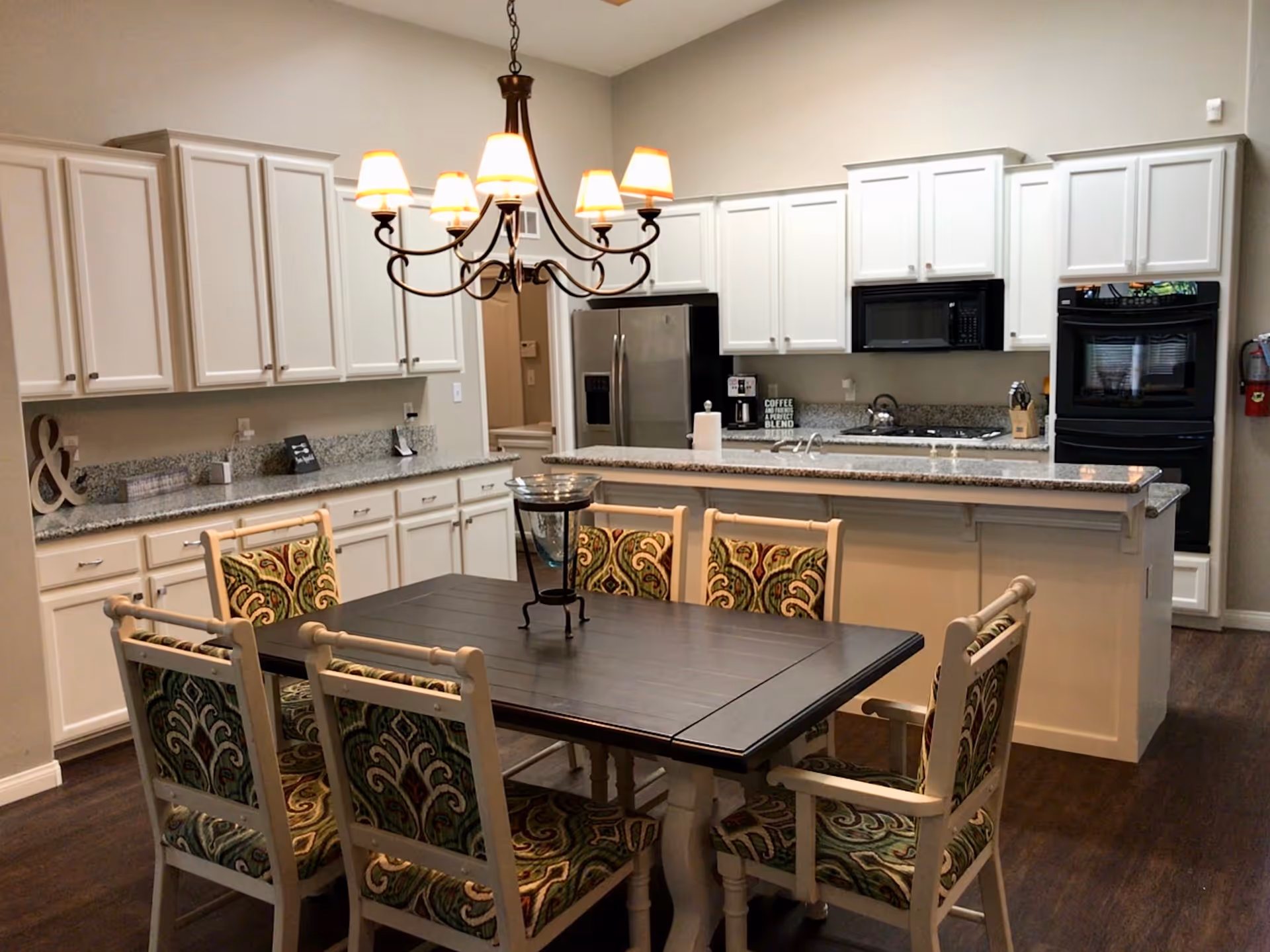 A bright kitchen and dining area with white cabinets, granite countertops, and stainless steel appliances. A dark wooden dining table with six patterned upholstered chairs is in the foreground, illuminated by a chandelier with six lampshades.